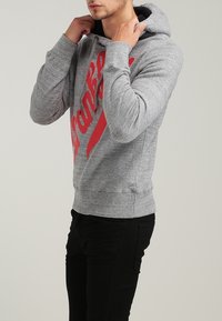 Grauer Hoodie mit einem roten Grafikdruck. Verfügt über eine Kapuze, gerippte Bündchen und einen gerippten Saum. Getragen mit schwarzen Hosen, leicht zur Seite gewandt.