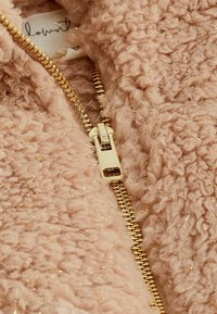 Gros plan d'une veste en tissu texturé rose clair avec des détails en fil doré et une fermeture éclair dorée partiellement ouverte.