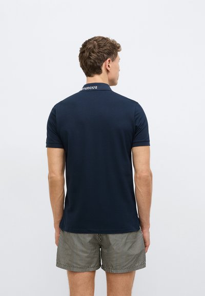 Tamsiai mėlyna polo marškinėliai su tekstūriniu dizainu, ant apykaklės balta siuvinėta logotipu, derinami su dryžuotomis šortais.