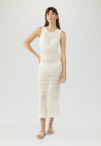 Robe tricot blanche sans manches avec un motif en losanges, longueur maxi, encolure côtelée, fentes sur les côtés, portée avec des sandales à fines lanières de couleur claire.