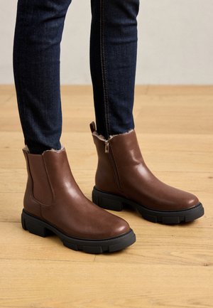 Brune læderankelstøvler med sidelommer og sorte chunky såler båret med mørkeblå skinny jeans på et trægulv.