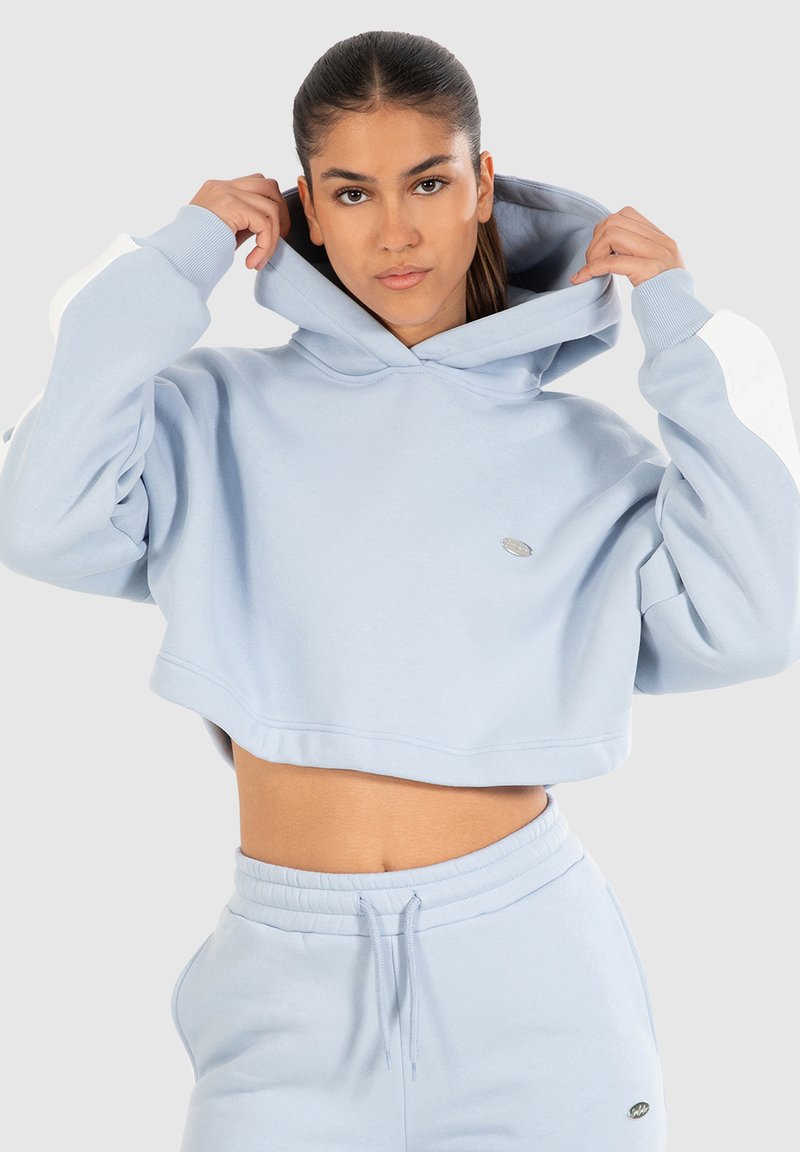Sudadera corta con capucha de color azul claro en tela suave, con acentos blancos en las mangas y un pequeño logo en la parte frontal. Complementa con joggers a juego.