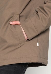 Khaki svrchní oděv s hladkou texturou, vybavený boční kapsou a prošívanými detaily. Malý štítek viditelný blízko lemu.