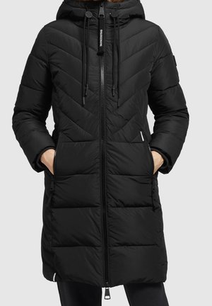 Czarna pikowana długa zimowa kurtka z kapturem, pełnym zamkiem z przodu, kieszeniami na zamek z boku oraz sznurkami przy dekolcie, noszona z rękami w kieszeniach.