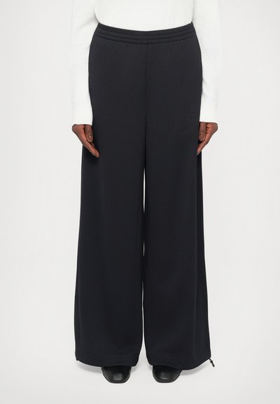 Personne portant un pantalon large texturé noir, un haut à manches longues blanc et des chaussures noires à bout carré, se tenant devant un fond blanc.