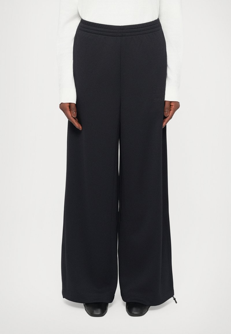 Personne portant un pantalon large texturé noir, un haut à manches longues blanc et des chaussures noires à bout carré, se tenant devant un fond blanc.