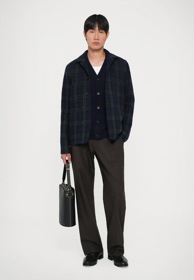 Giovane uomo che indossa una giacca a scacchi scura, un cardigan blu navy, una camicia bianca, pantaloni marroni e scarpe nere, mentre porta una valigetta in pelle nera.