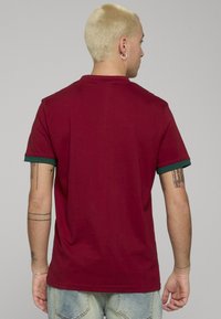 Burgunderfarget T-skjorte med grønn kant på kragen, korte ermer og en løs passform. Laget av myk bomull med minimal tekstur og detaljer.