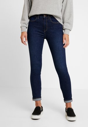 Personne portant un jean skinny bleu foncé roulé aux chevilles, un haut à manches longues gris et des chaussures slip-on noires avec des semelles blanches.