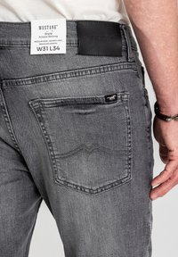 Szare dżinsy z denimu o wąskim kroju, średnim stanie, z czarną skórzaną naszywką w pasie oraz wzorem na tylniej kieszeni.