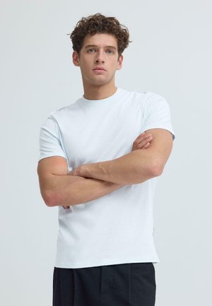 Junger Mann mit lockigem Haar, der ein schlichtes weißes T-Shirt und schwarze Hose trägt, steht mit verschränkten Armen vor einem hellgrauen Hintergrund.