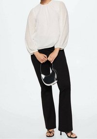 Blusa bianca strutturata con maniche a palloncino, abbinata a pantaloni neri aderenti e sandali neri con cinturini. Tiene in mano una piccola borsa nera a forma curva.