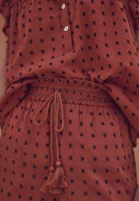 Haut en terracotta avec des motifs carrés noirs, doté d'une patte de boutonnage et d'une taille smockée avec un cordon de serrage et des accents de gland.
