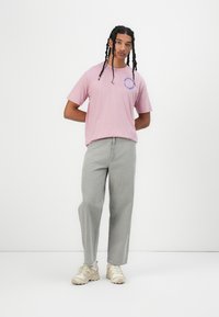 Pink cotton t-shirt with logo graphic in blue, paired with light gray wide-leg jeans and beige sneakers, worn by a person with braided hair.