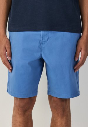 Mand iført enkle lyseblå shorts til knæet og en mørk marineblå skjorte, stående med afslappede arme langs siderne mod en neutral baggrund.