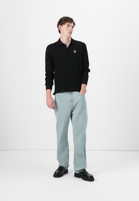Musta pikade varrukatega polo särk, millel on rindkeres logo, koos helesiniste vabade jeanidega ja mustade kingadega, seistes ühetoonilise tausta ees.
