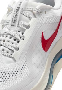 Baskets de sport blanches avec une tige en maille, un swoosh rouge de Nike et des accents texturés. Elles présentent des lacets gris et une unité de coussin d'air visible.