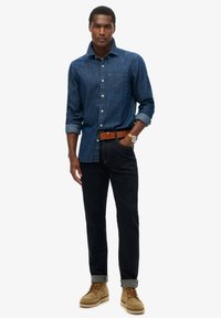 Camisa de mezclilla de botones en azul oscuro, de manga larga, con un bolsillo en el pecho. Combinada con pantalones oscuros, botas beige y un cinturón marrón.