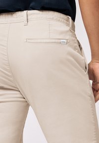 Beige cotton trousers featuring a flat waistband, single back pocket with a label, and side pockets. Smooth texture with a fitted design.
