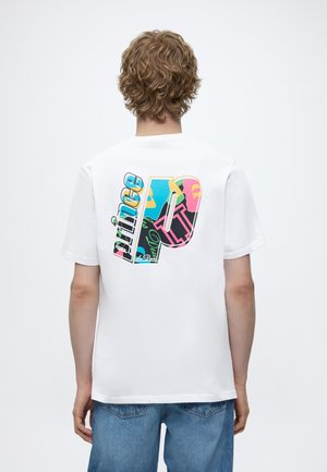 Personne aux cheveux bouclés, vue de dos, portant un T-shirt blanc avec un grand « P » coloré et le texte « prince » au dos.