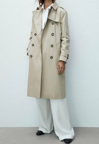 Femme portant un trench-coat beige à double boutonnage sur un pantalon blanc à jambes larges et des chaussures noires, debout contre un mur clair uni.