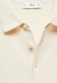 Kremowa polo koszulka z teksturowanym splotem, klasycznym kołnierzykiem i trzema przyciskami przypominającymi perły z przodu. Widoczna metka marki.