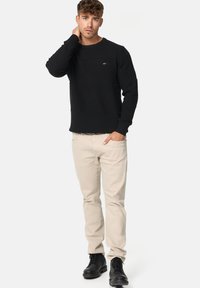 Zwarte gestructureerde trui met een geribbeld patroon, beige broek en zwarte veterschoenen. Model poseert met één hand op de nek, wat de casual stijl benadrukt.