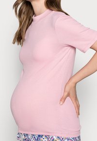 Světle růžový žebrovaný top s krátkým rukávem a vysokým límcem, nošený přes křivkový střih. Vyznačuje se hladkou texturou a výraznou látkou.