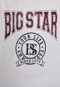 Valge T-särk, millel on tumepunased kulunud tähed "BIG STAR" ja must ümmargune logo, millel on tekst "YOUR LIFE SINCE 1979" ja "FS" sees.