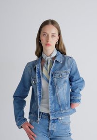 Denim jakker klassikalise lõikega, helesinine värv, kaks rinna taskut ja metallnupud. Kantud valge särgi ja heledat mustriga salliga.