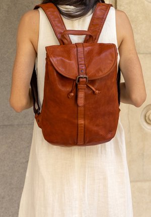 Brown leather backpack with a flap closure, drawstring detail, and adjustable shoulder straps. Features a textured surface and subtle stitching.