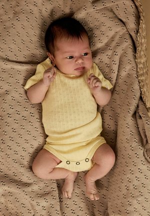 Noworodek leży na beżowym, dzierganym kocu, ubrany jest w blady żółty body, spogląda lekko na bok ze ściśniętymi pięściami.