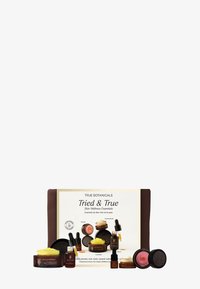 Set von Hautpflegeprodukten in einem braunen Beutel, bestehend aus verschiedenen kleinen Gläsern und Flaschen mit Etiketten, darunter bernsteinfarbenes Glas, gelbe Creme und matte schwarze Akzente.