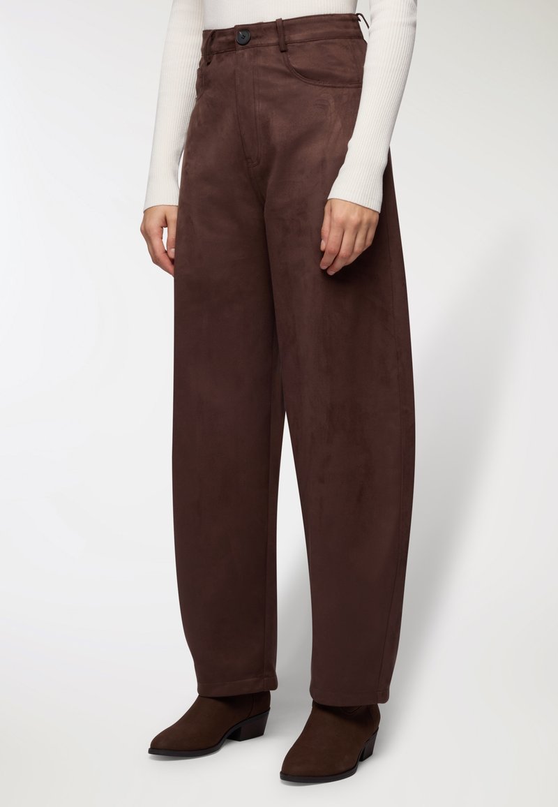 Persona che indossa pantaloni larghi a vita alta di colore marrone scuro e stivaletti con tacco marrone scuro, abbinati a un top a maniche lunghe bianco a coste.