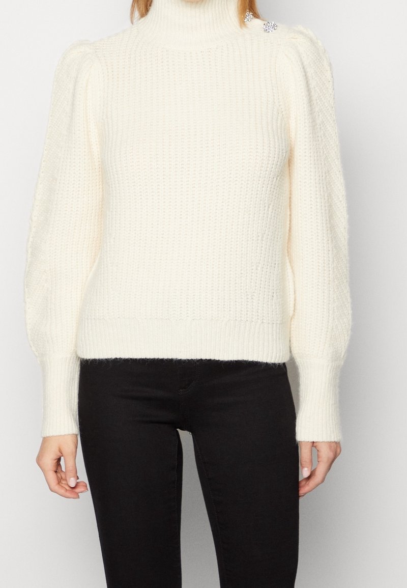 Maglione a collo alto crema in maglia spessa con superficie texturizzata, spalle a sbuffo, polsini a coste e un accento decorativo vicino alla spalla.