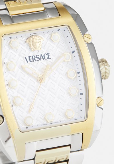 Relógio Versace com caixa metálica quadrada em prata e ouro, mostrador texturizado branco apresentando marcadores de horas hexagonais e logotipo. Detalhes em ouro nos ponteiros.