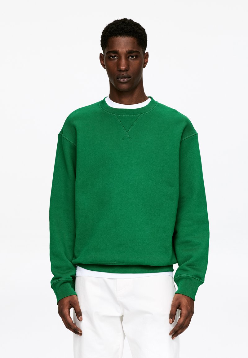 Sweat-shirt vert en tissu doux. Présente un col rond, des poignets côtelés et une coupe décontractée. Porté par-dessus une chemise blanche.