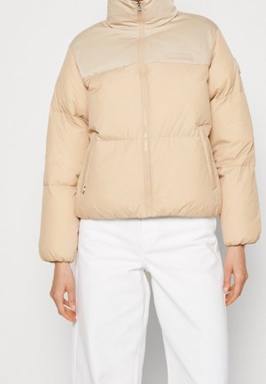 Beige cropped donzen jack met hoge kraag en ritssluiting aan de voorkant, gedragen met een witte broek, alleen torso en armen zichtbaar.