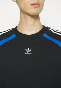 Černé tričko Adidas s modrými akcenty na ramenou, žebrovaným kulatým výstřihem a bílým logem na hrudi. Měkká, lehká tkanina.