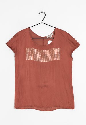 Blouse à manches courtes couleur rouille avec un col arrondi, présentant un motif horizontal de petites découpes carrées blanches sur la poitrine, suspendue à un cintre noir.