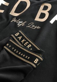 Czarna tkanina z dużymi metalicznymi złotymi literami. Zawiera metki z napisem "TED BAKER" w złocie na czarnym tle z dekoracyjnymi paskami.