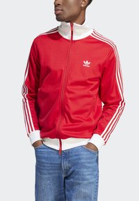 Red zip-up jacket with white collar and cuffs, featuring three white stripes on each sleeve. Small white logo on the left chest.