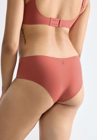 Vrouw in een naadloze dusty rose beha en bijpassende high-waist onderbroek, met haar rug naar de kijker gekeerd en een tatoeage aan de zijkant.