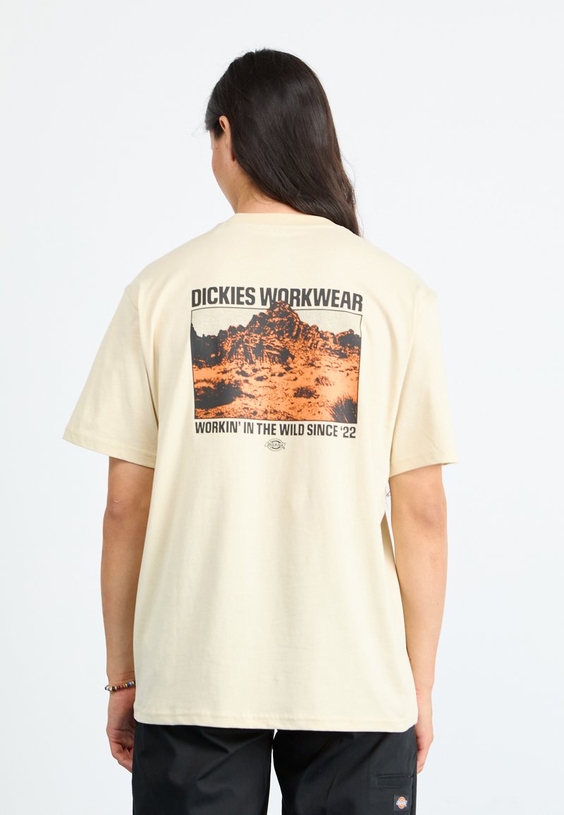 Lys beige t-shirt med grafisk tryk på ryggen. Har fed tekst og et bjerglandskab i orange og sort. Korte ærmer.