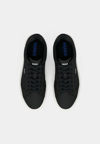 Μαύρα αθλητικά παπούτσια με επάνω μέρος από λείο ύφασμα, με λευκά κορδόνια και καουτσούκ σόλα. Το "HUGO" τυπωμένο με μπλε χρώμα στις εσωτερικές σόλες και τις πλαϊνές πλευρές.