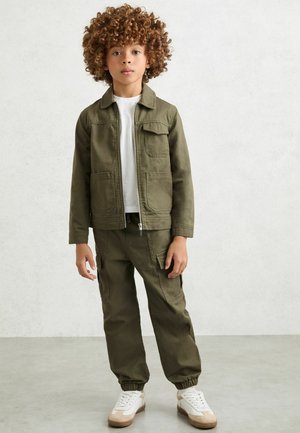 Copil cu păr creț, purtând o jachetă și pantaloni verde măsliniu, cămașă albă și adidași albi, stând în fața unui perete simplu, deschis la culoare.