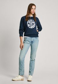 Felpa blu navy con logo circolare bianco, abbinata a jeans blu chiaro. Modello in piedi con sneakers bianche, su uno sfondo neutro.