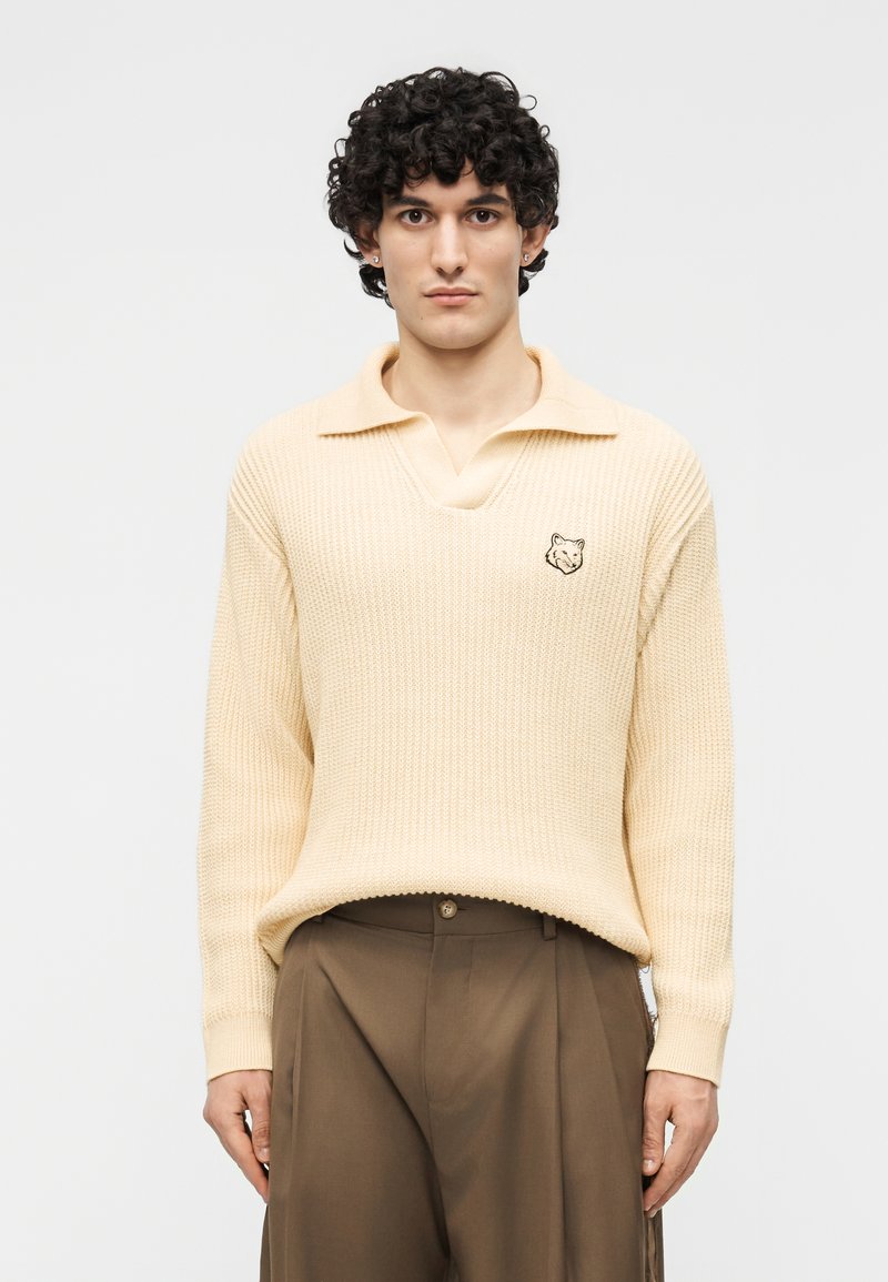 Jeune homme aux cheveux bouclés portant un pull crème en maille avec un écusson de loup et un pantalon marron ample, debout devant un fond uni.