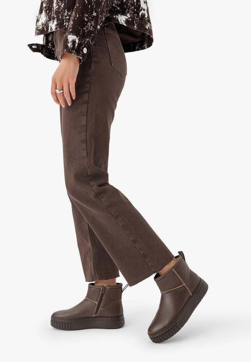 Person, der braune gerade geschnittene Hosen und dunkelbraune Ankle Boots mit seitlichem Reißverschluss trägt, geht vor einem schlichten Hintergrund.