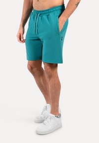 Pantalones cortos de color aguamarina con cintura elástica, cordón ajustable y bolsillos laterales. Están hechos de una tela suave y cuentan con un pequeño parche de logo en la pierna.